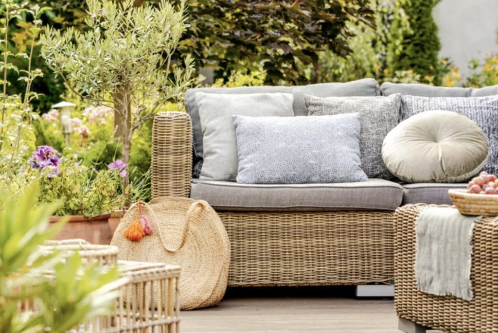 Light brown wicker furniture with gray cushions and lots of gray and white pillows piled ont he couch in front of a lushly green backyard with a few purple flowers peeking out of a pot.