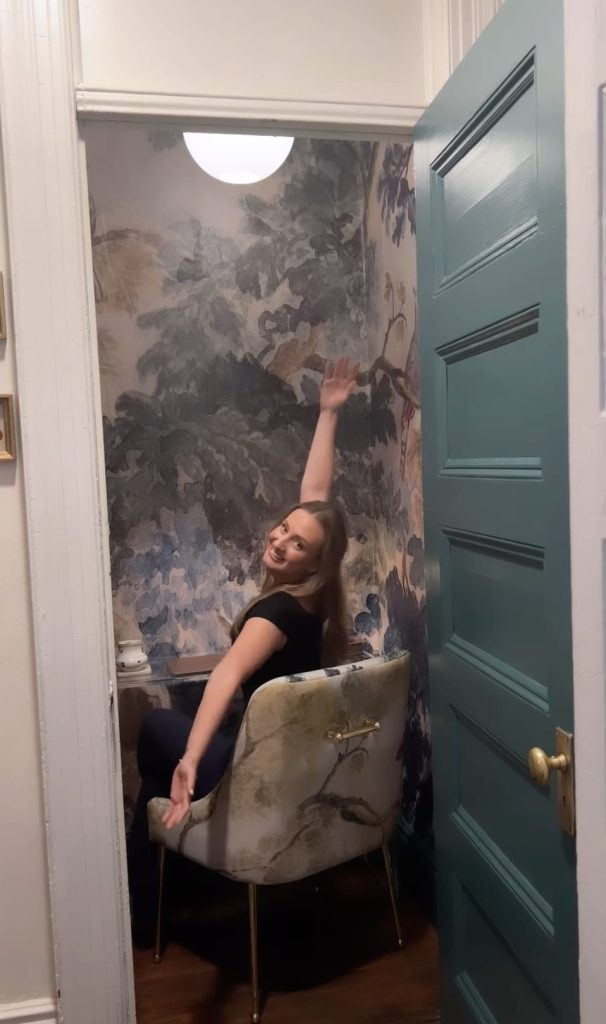 A young woman sits at a desk with her arms spread wide sharing the beautifully redone room. There is a teal colored door and beautifdul watercolor wallpaper with matching chair in the room