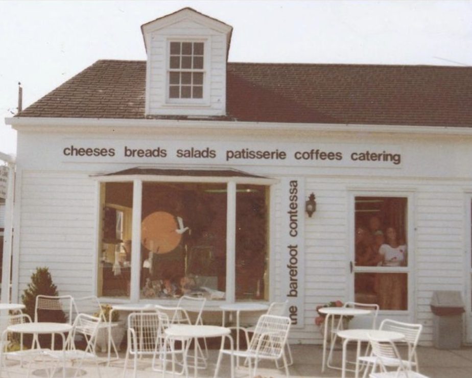 Storefront of Ina Garten's store Barefoot Contessa which was opened 45 years ago. Storefront is white clapboard with lowercase black letters spelling "the barefoot contessa, cheeses, breads, salads, patisserie, coffees, catering. There are white chairs and tables in front of the store