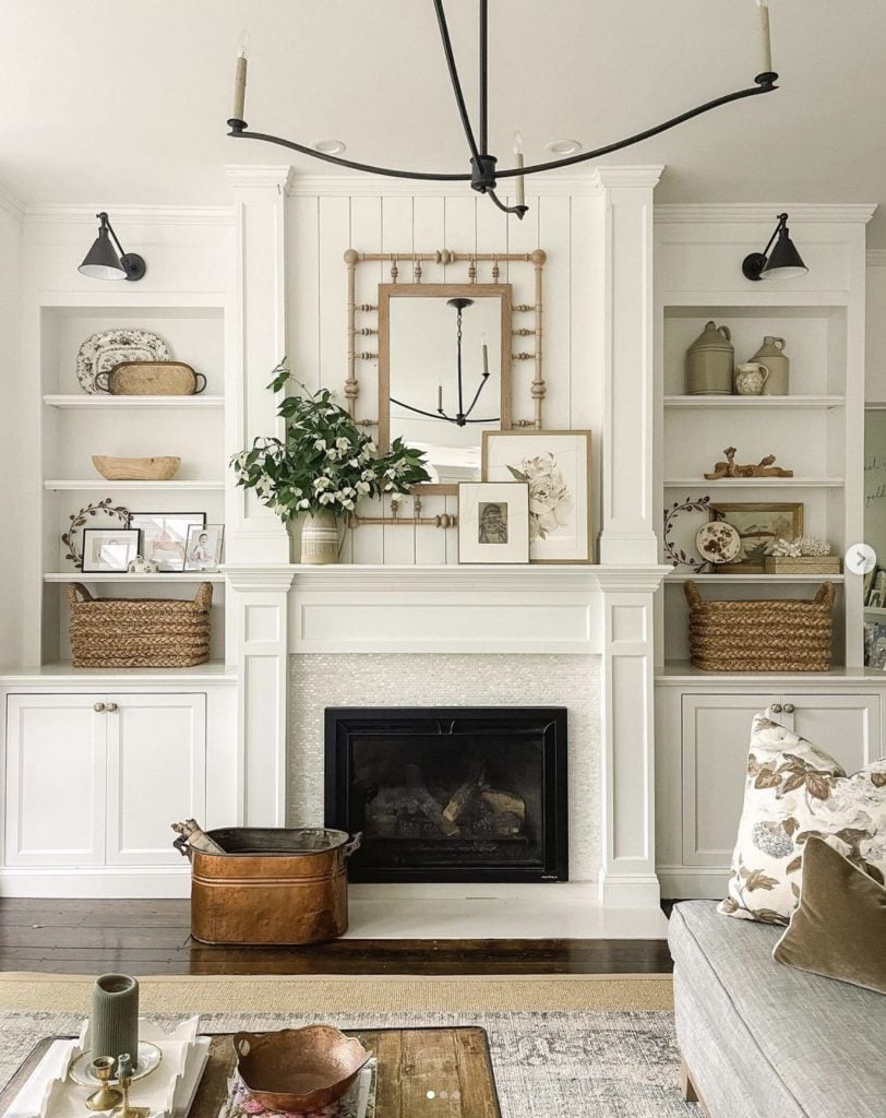 A closeup photo of a livingroom fireplace flanked by bookcases on each side. The room is decorated in shades of white and tan. On the mantel is a bouquet of dogwood in a pottery vase. A mirror made from wood spindles over the fireplace reflects a slim iron arm chandelier. The bookcases are artfully decorated with baskets, pottery and paintings/pictures in frames all in the same white/tan tones. A copper bucket with pieces of wood is at the hearth of the fireplace.