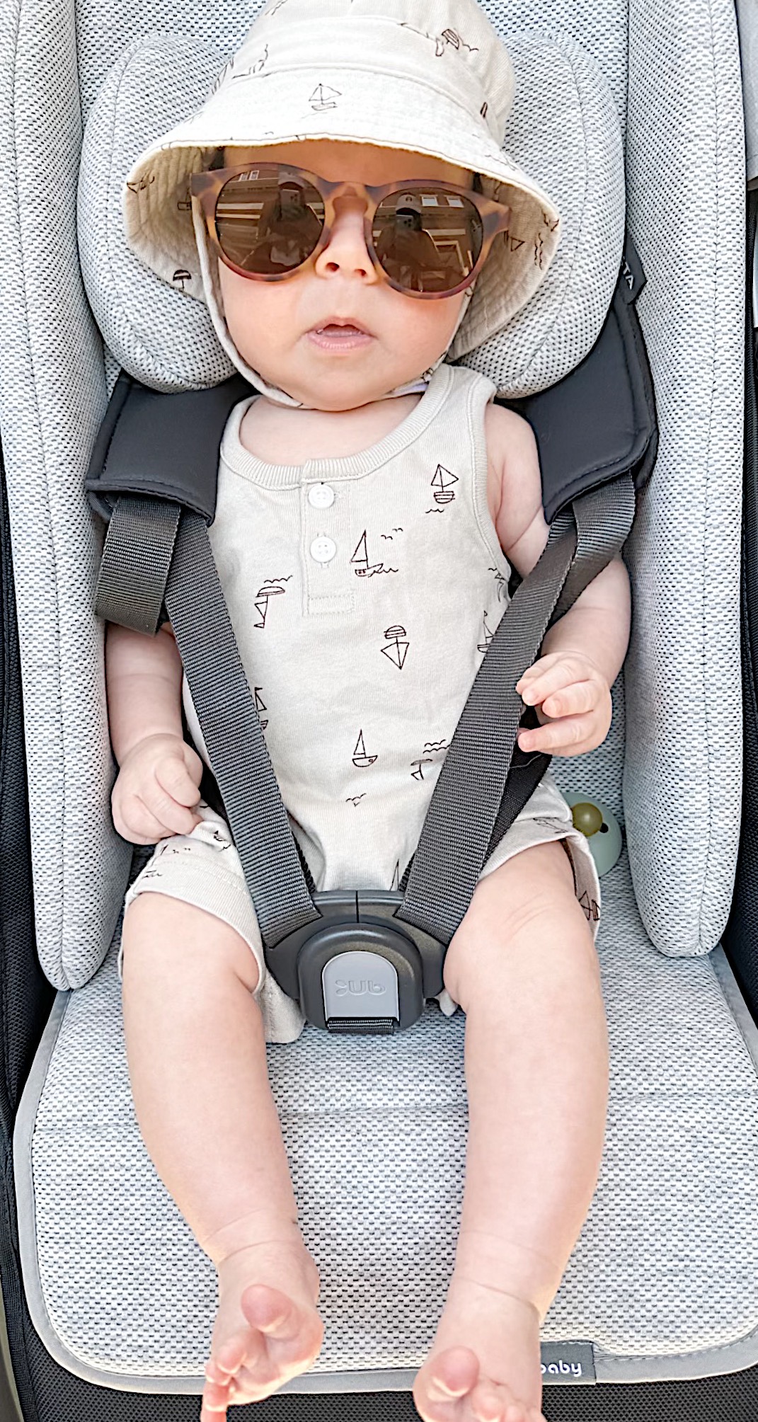 Our grandson in his carseat ready for the beach in a onesie, sunglasses, and a sun hat.