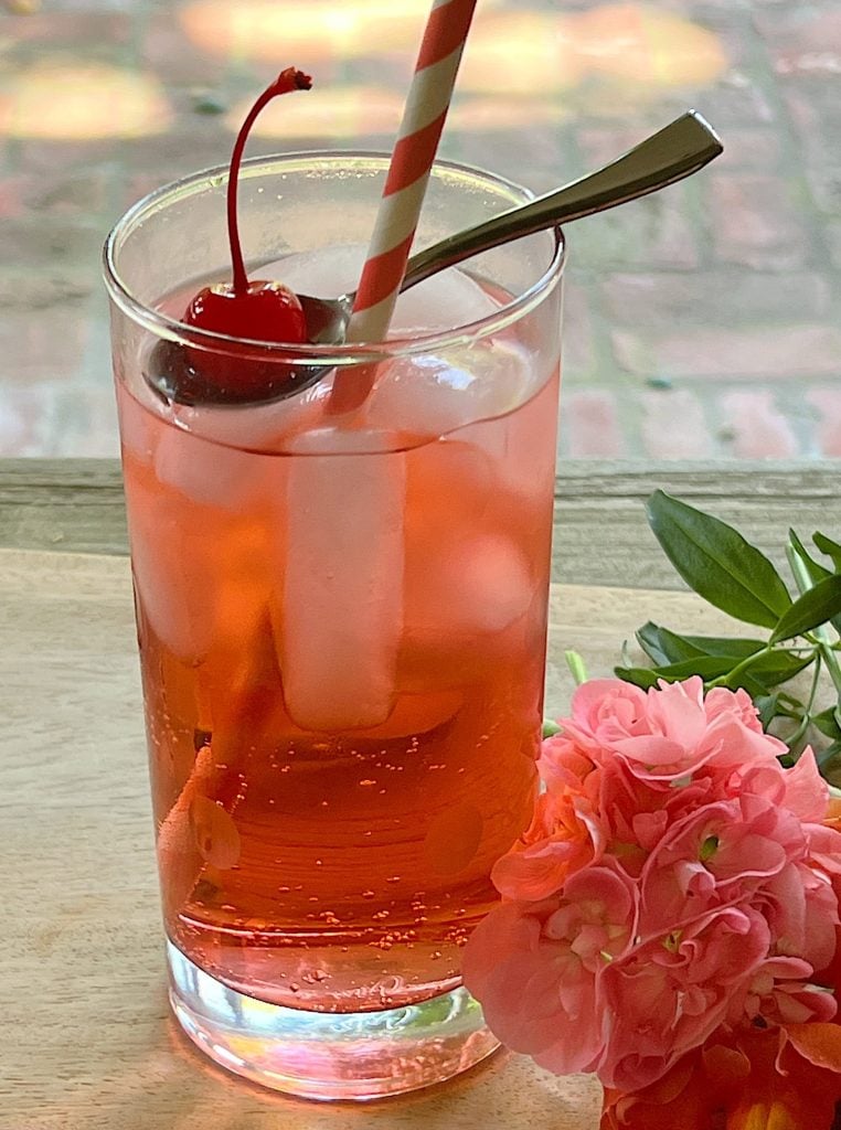 A freshly made Shirley temple Refreshing Summer Mocktail in a glass with a paper straw and a marachino cherry.
