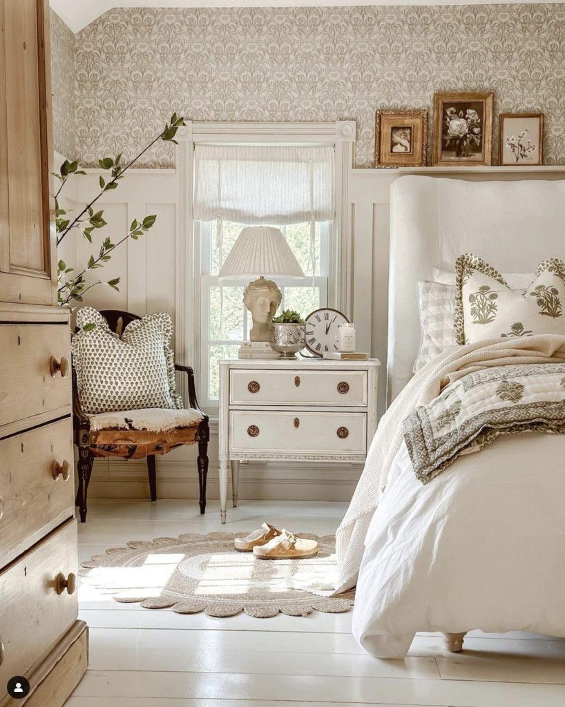 A beautiful bedroom all in white and beige colors. A light wood dresser is in the foreground, wood floors are painted white and walls have white wainscoting, a desconstructed chair sites in the corner next to a white side table filled with a cement but lamp, a silver pot and antique clock. The bed frame is covered in white linen and bedding is white and being. There are three gold froms leaning against the wall at the top of the wainscoting with netral floral paintings.