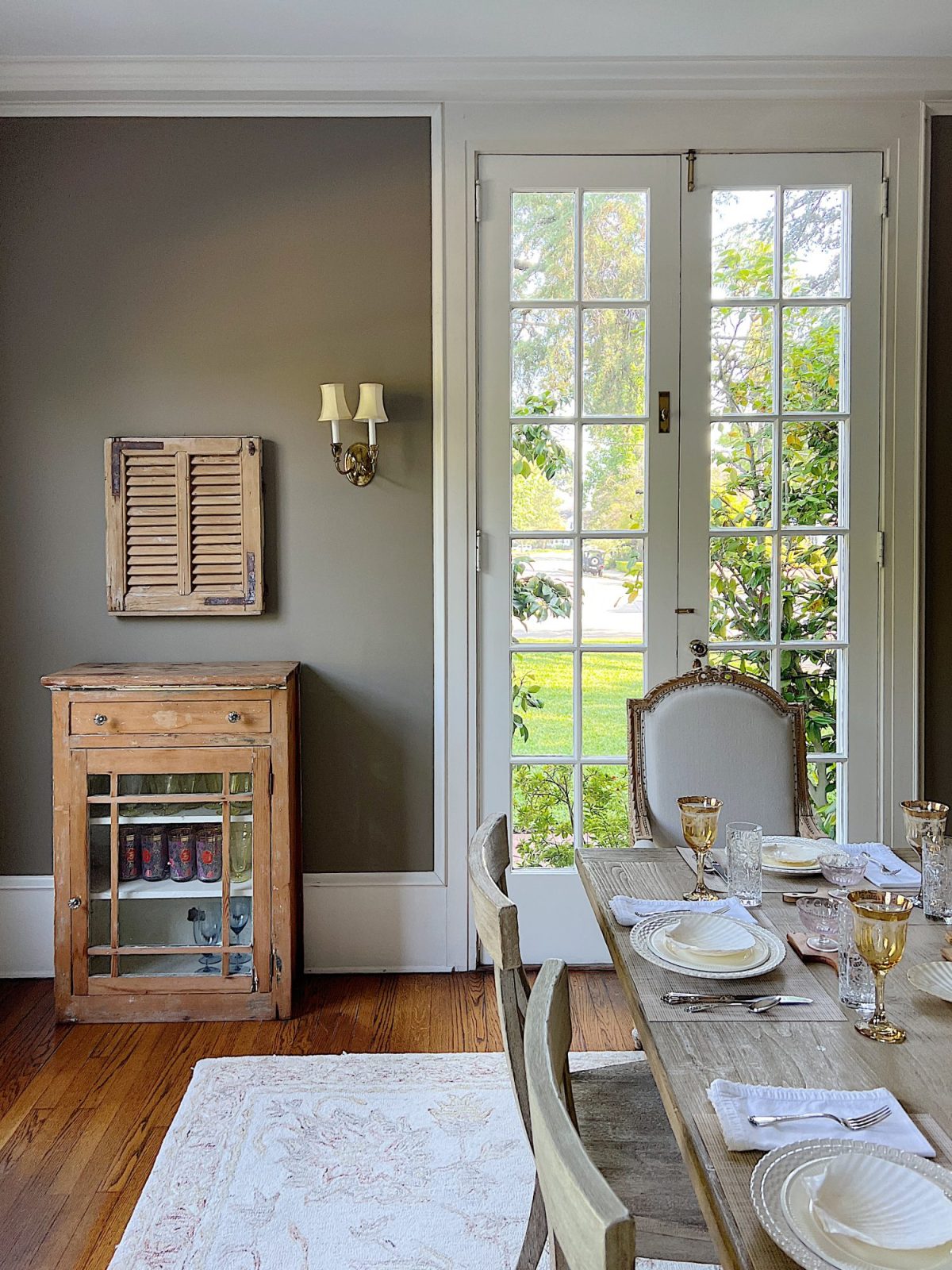 A dining room with light brown walls, white trim, a vintage chest and french shutter, and a farmhouse wood dining room table.