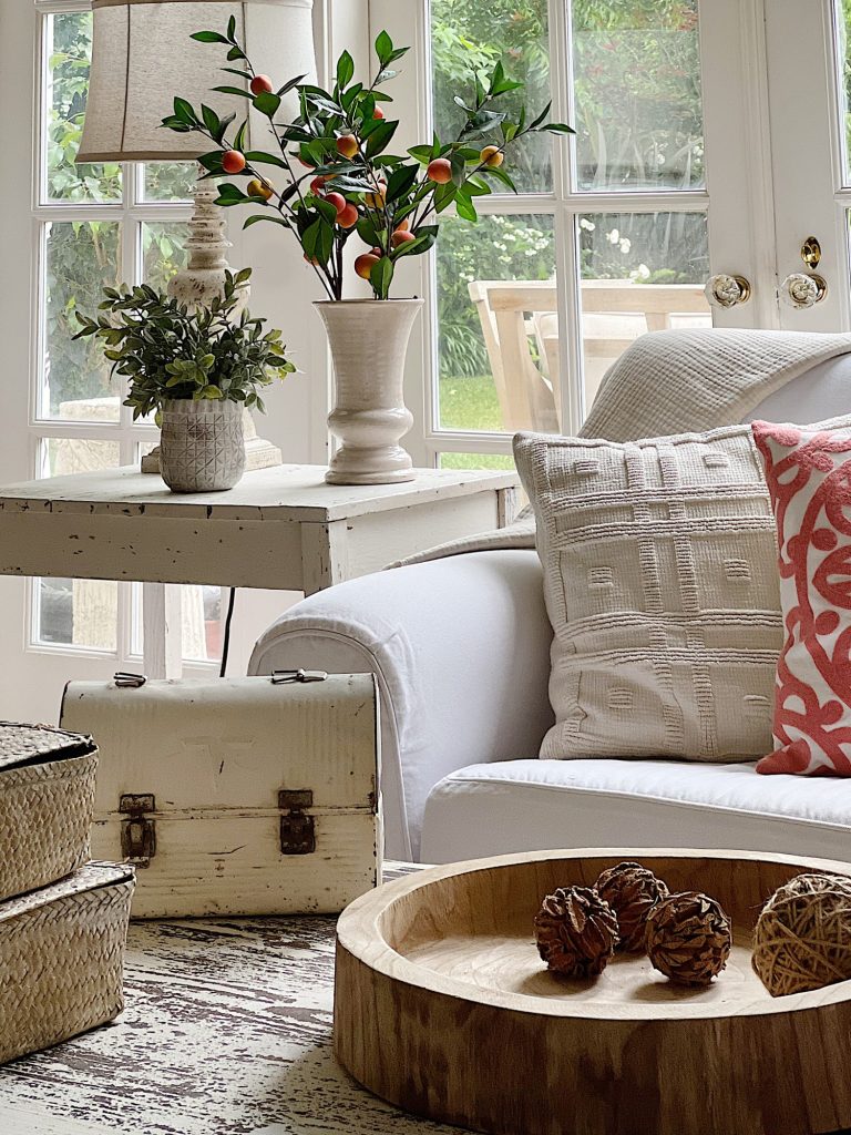 A white couch and assorted pillows and a vintage hanging chandelier are also in the family room. There are peach blossoms in a vase and various neutral decor items on the coffee table.