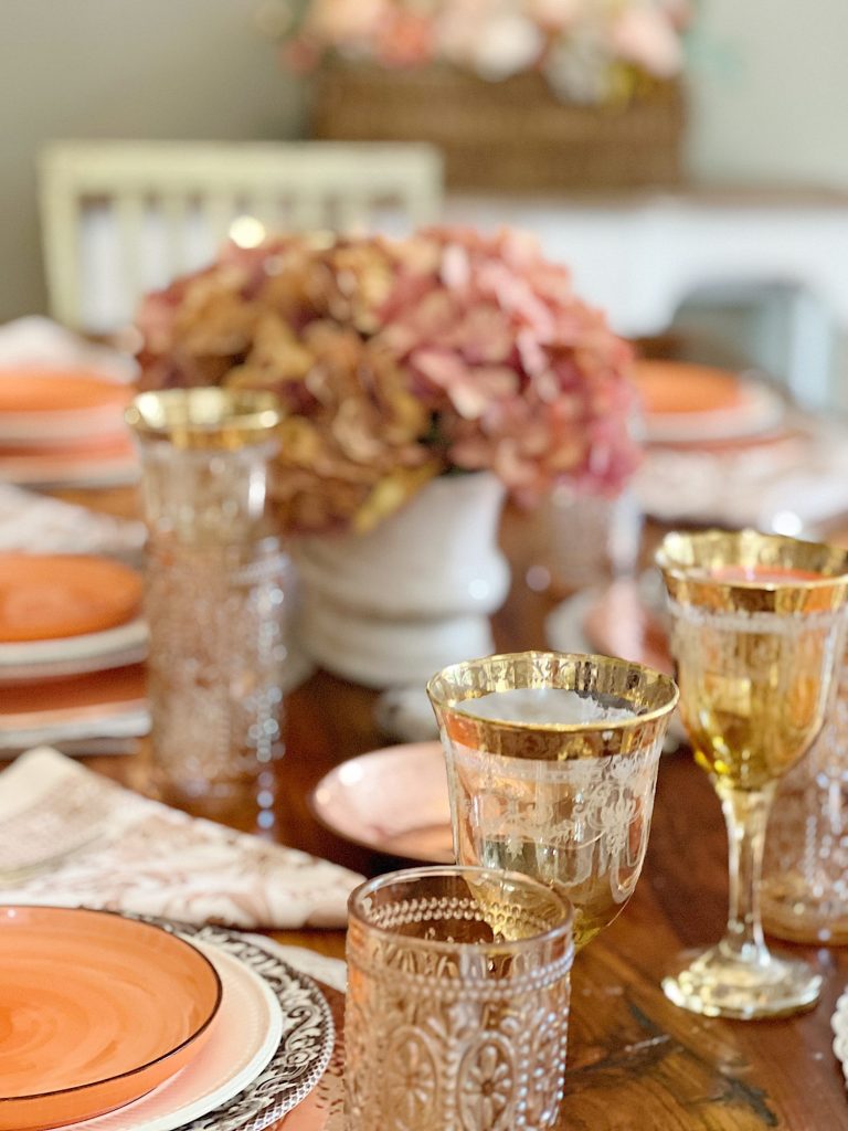 A dining table in a family room with white, brown, and orange plates, copper accents, amber colored glasses, and a faux pinkish orange hydrangea centerpiece