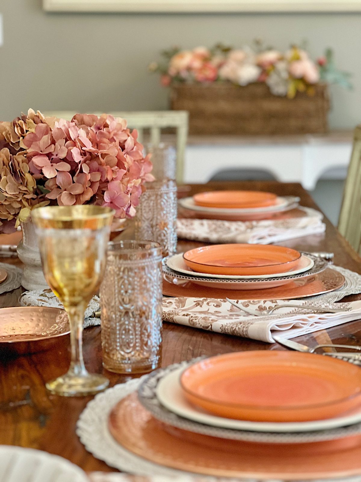 A dining table in a family room with white, brown, and orange plates, copper accents, amber colored glasses, and a faux pinkish orange hydrangea centerpiece