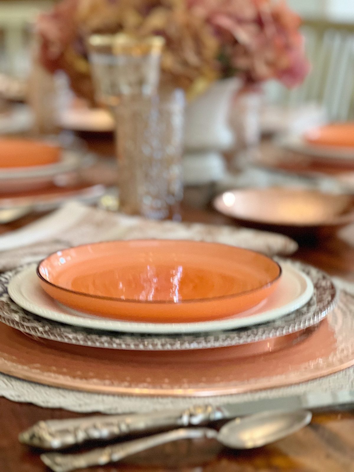 A dining table in a family room with white, brown, and orange plates, copper accents, amber colored glasses, and a faux pinkish orange hydrangea centerpiece