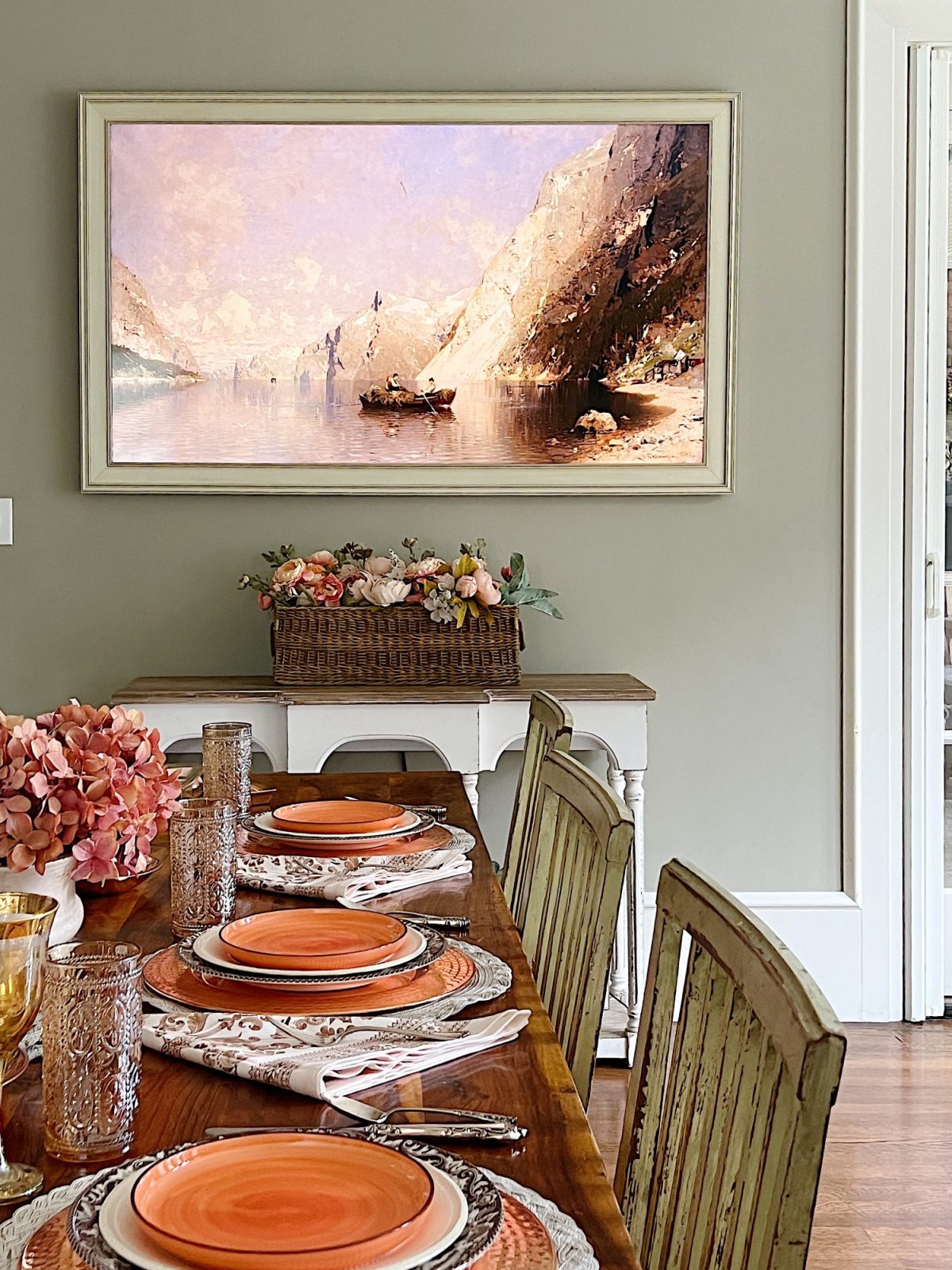 A dining table in a family room with white, brown, and orange plates, copper accents, amber colored glasses, and a faux pinkish orange hydrangea centerpiece
