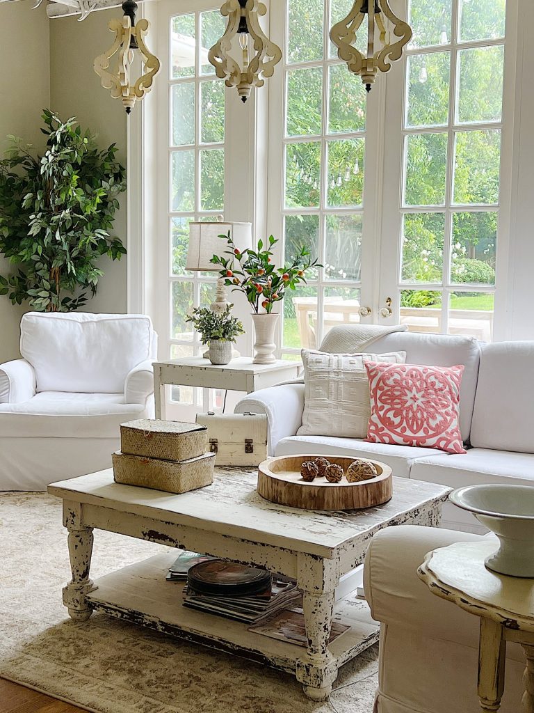 A white couch and assorted pillows and a vintage hanging chandelier are also in the family room. There are peach blossoms in a vase and various neutral decor items on the coffee table.