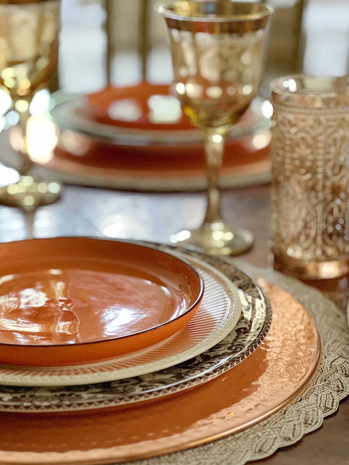 A dining table in a family room with white, brown, and orange plates, copper accents, amber colored glasses, and a faux pinkish orange hydrangea centerpiece