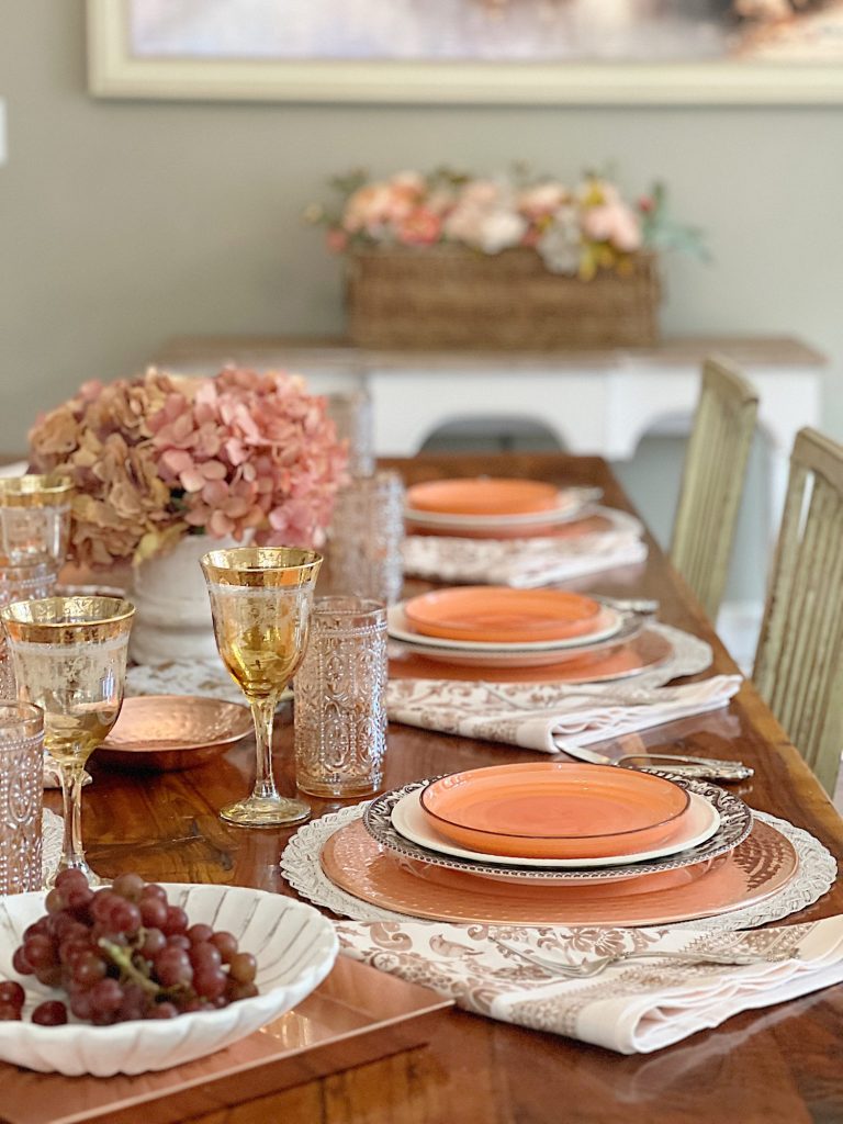 A dining table in a family room with white, brown, and orange plates, copper accents, amber colored glasses, and a faux pinkish orange hydrangea centerpiece