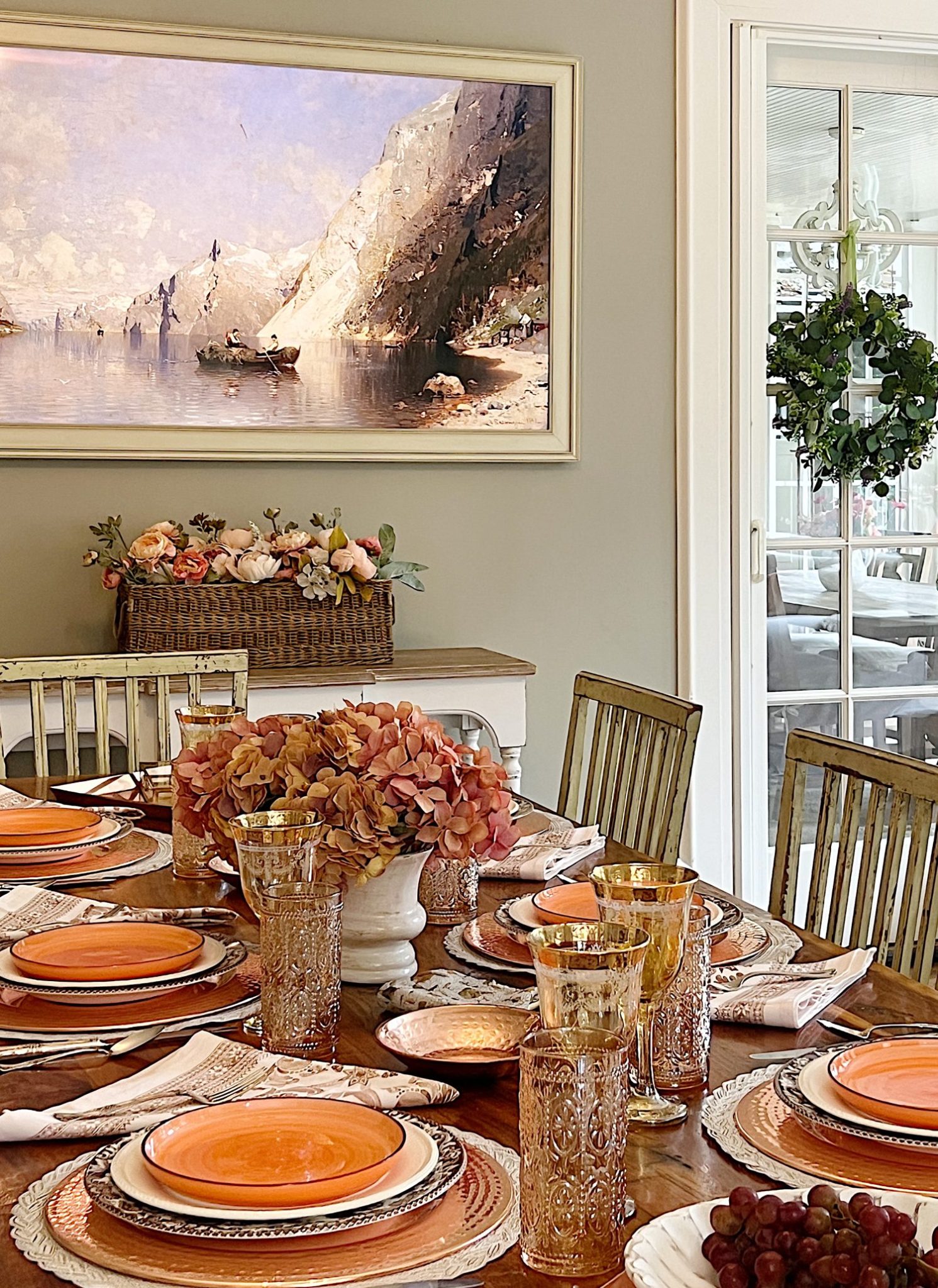 A dining table in a family room with white, brown, and orange plates, copper accents, amber colored glasses, and a faux pinkish orange hydrangea centerpiece
