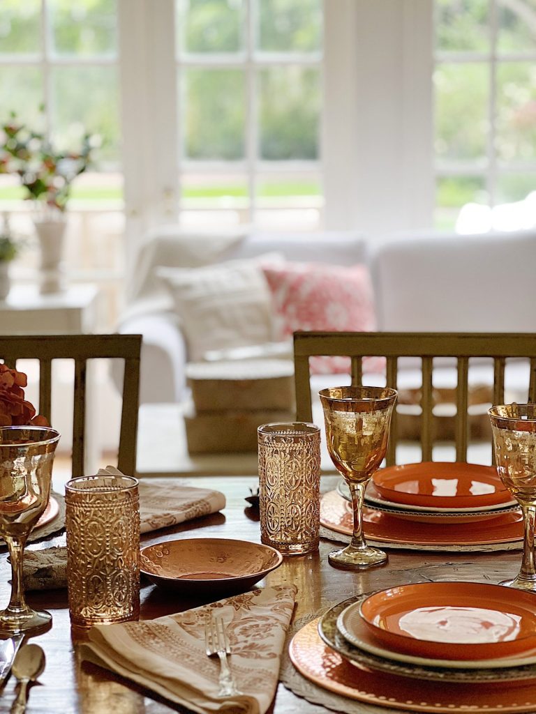 A dining table in a family room with white, brown, and orange plates, copper accents, amber colored glasses, and a faux pinkish orange hydrangea centerpiece. A white couch and assorted pillows and a vintage hanging chandelier are also in the family room.