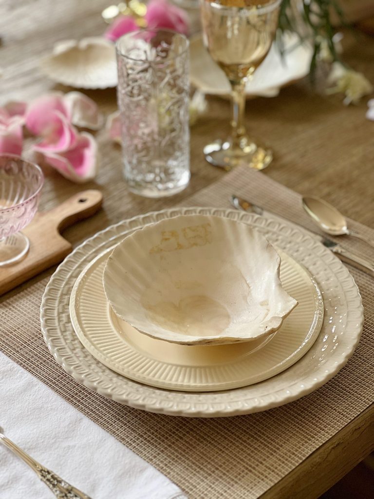 A summer table set with off white dishes, amber and clear glassware, and fresh pink and white flowers in an oval ceramic vase.