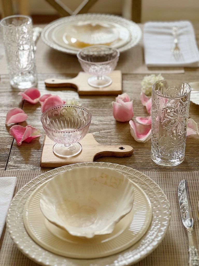 A summer table set with off white dishes, amber and clear glassware, and fresh pink and white flowers in an oval ceramic vase.