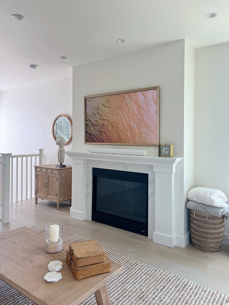 The living area at our beach house with a large fireplace, a frame tv, a window overlooking the ocean, and a couch, rug, coffee table and chair from Pottery Barn. The vintage cabinet, marble bust, and round rattan mirror are at the top of the staircase.