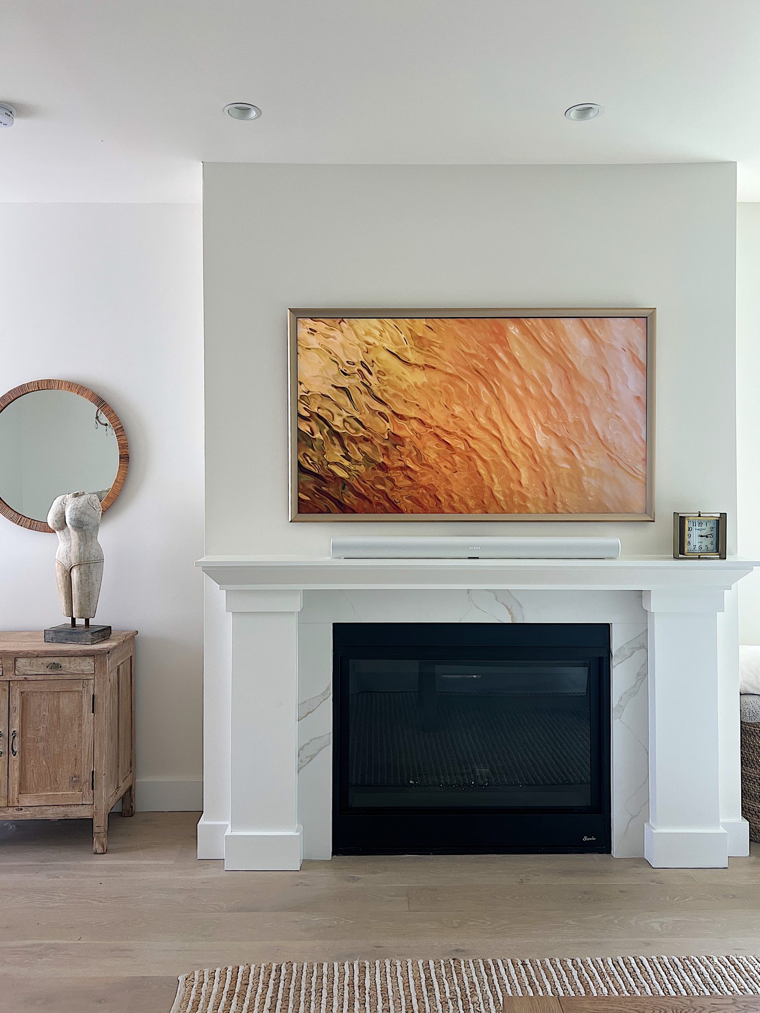 The living area at our beach house with a large fireplace, a frame tv, a window overlooking the ocean, and a couch, rug, coffee table and chair from Pottery Barn. The vintage cabinet, marble bust, and round rattan mirror are at the top of the staircase.