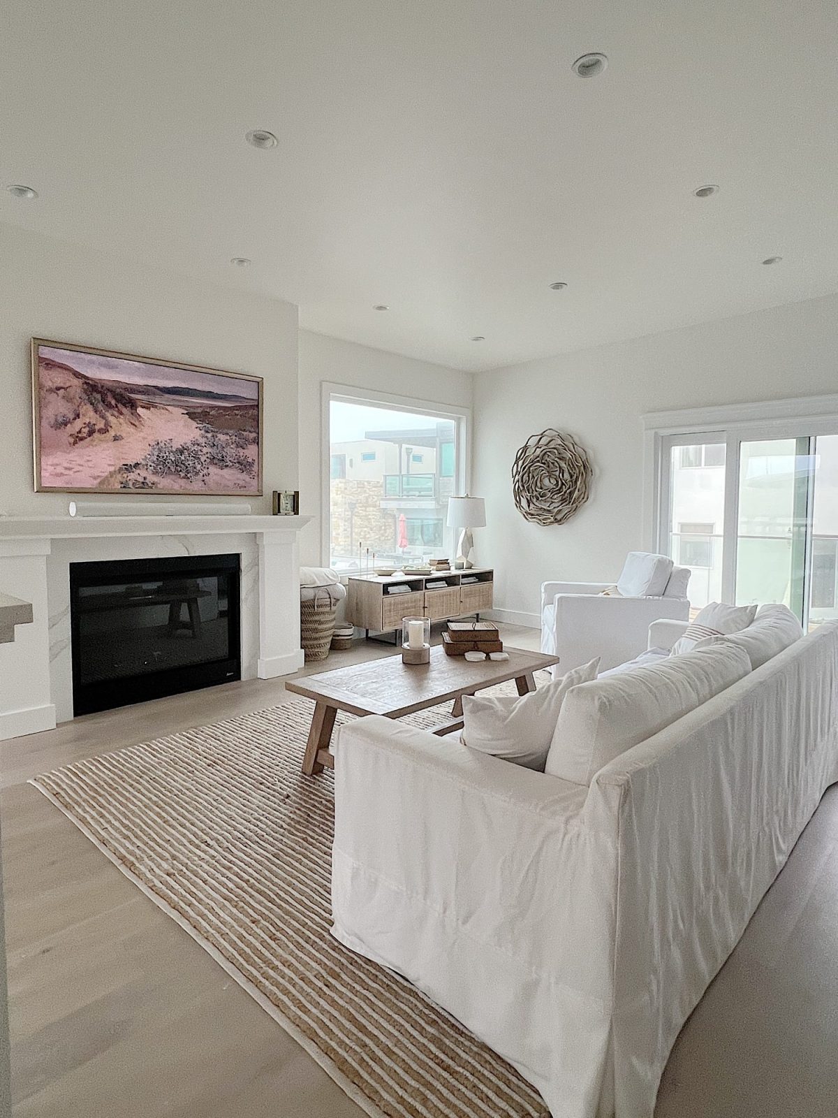 The living area at our beach house with a large fireplace, a frame tv, a window overlooking the ocean, and a couch, rug, coffee table and chair from Pottery Barn. The vintage cabinet, marble bust, and round rattan mirror are at the top of the staircase.