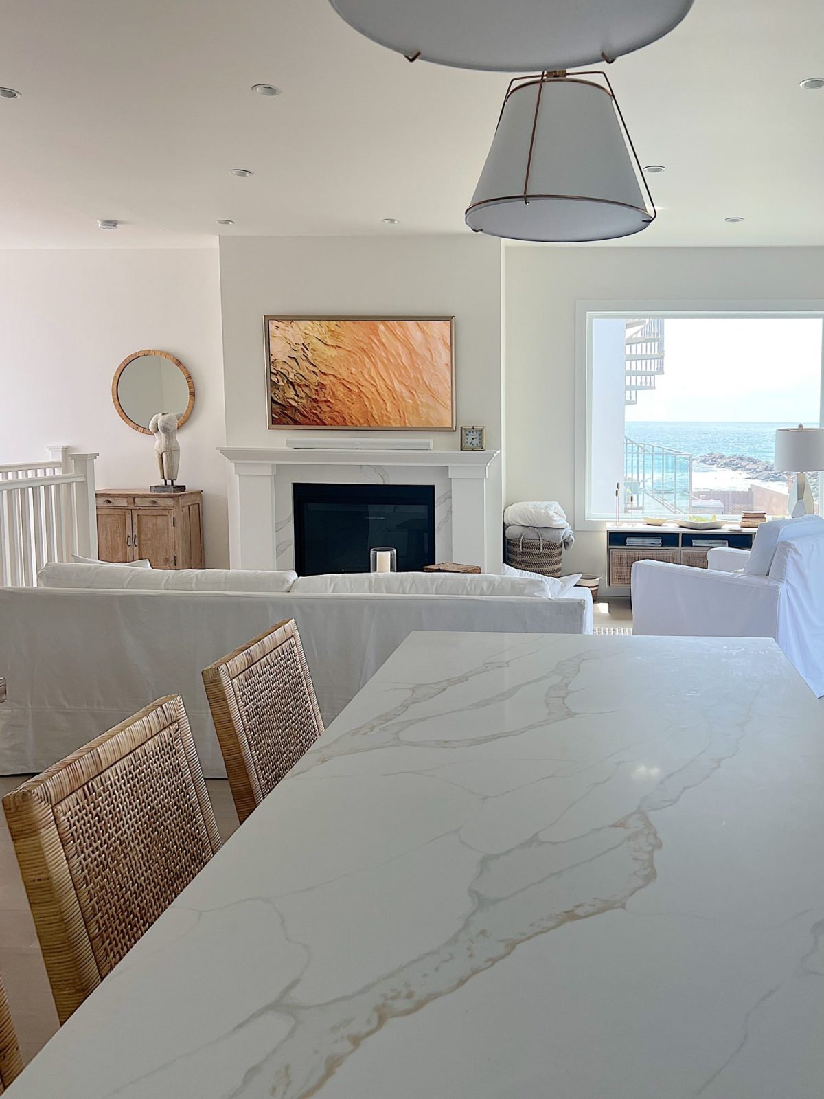 The living area at our beach house with a large fireplace, a frame tv, a window overlooking the ocean, and a couch, rug, coffee table and chair from Pottery Barn. The vintage cabinet, marble bust, and round rattan mirror are at the top of the staircase.