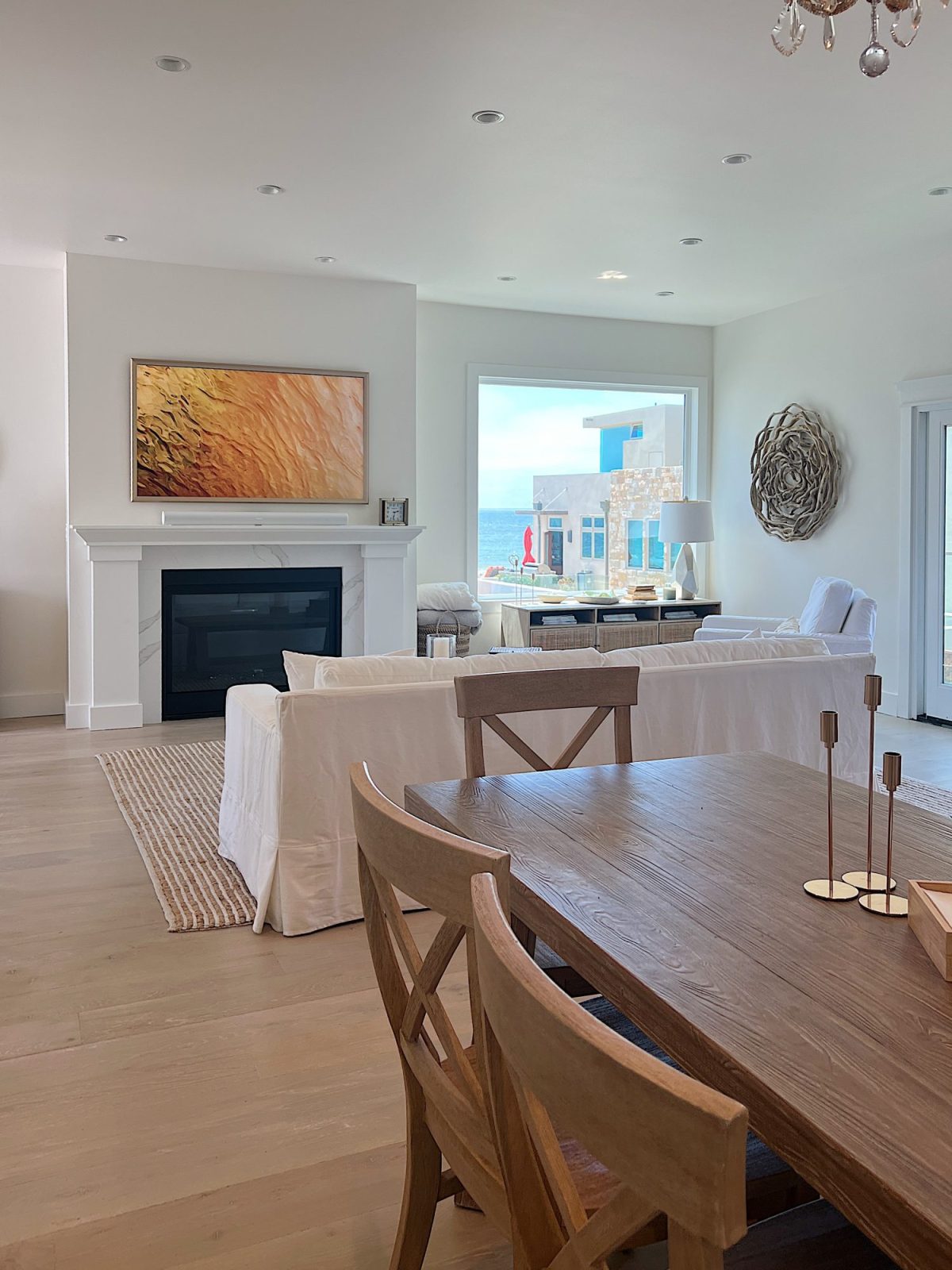 The living area at our beach house with a large fireplace, a frame tv, a window overlooking the ocean, and a couch, rug, coffee table and chair from Pottery Barn. The vintage cabinet, marble bust, and round rattan mirror are at the top of the staircase.