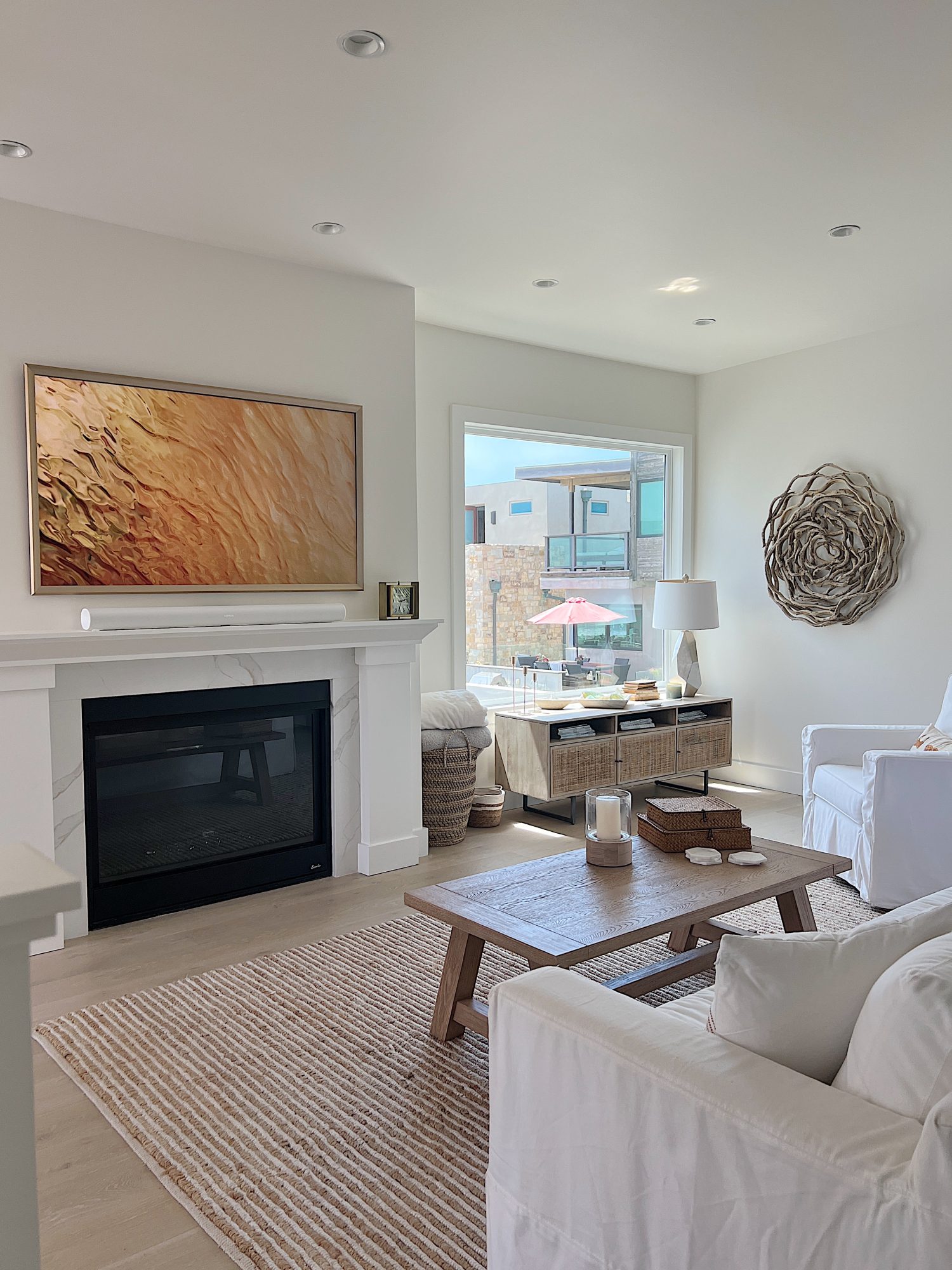 The living area at our beach house with a large fireplace, a frame tv, a window overlooking the ocean, and a couch, rug, coffee table and chair from Pottery Barn. The vintage cabinet, marble bust, and round rattan mirror are at the top of the staircase.