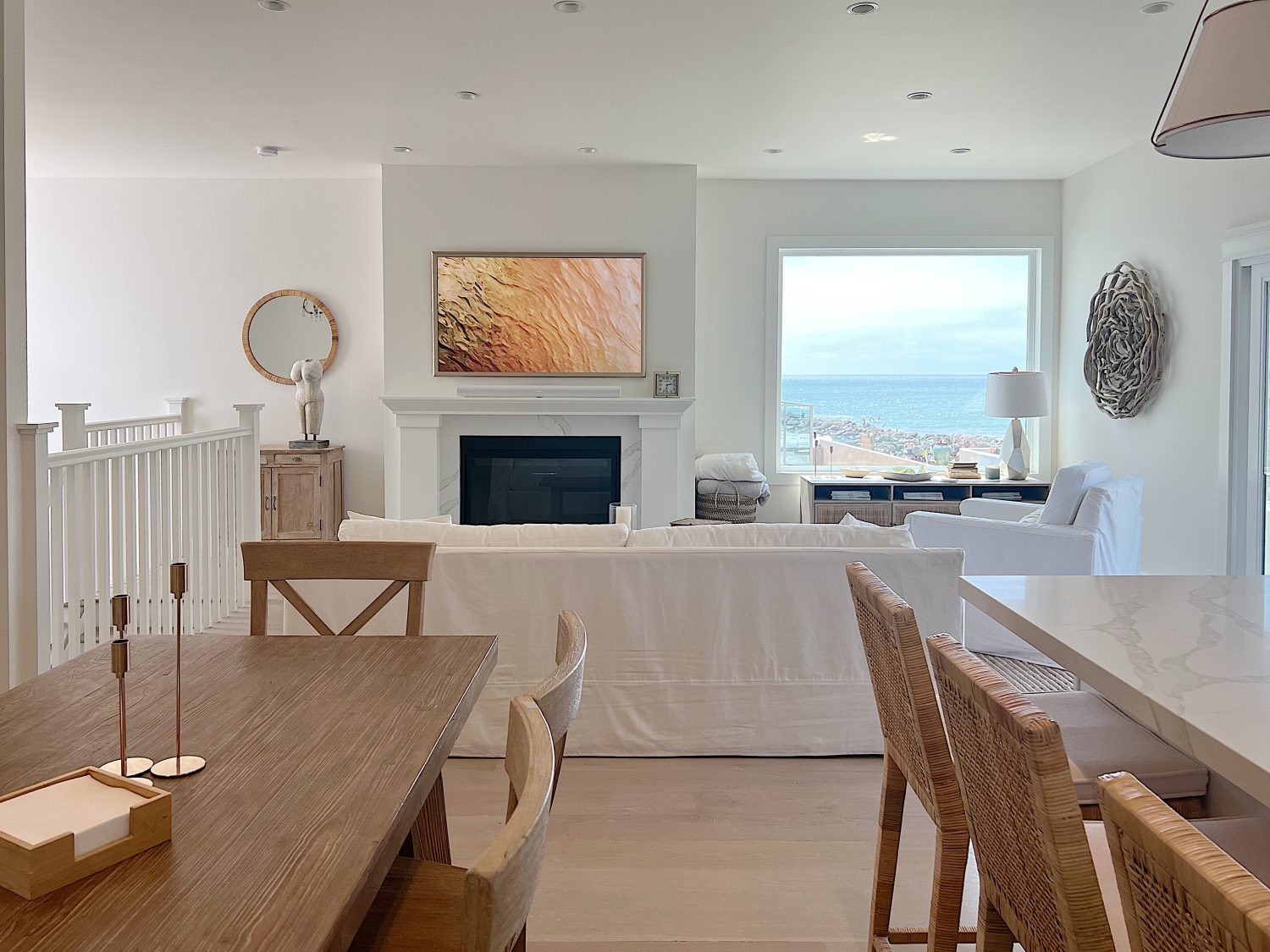 The living area at our beach house with a large fireplace, a frame tv, a window overlooking the ocean, and a couch, rug, coffee table and chair from Pottery Barn. The vintage cabinet, marble bust, and round rattan mirror are at the top of the staircase.