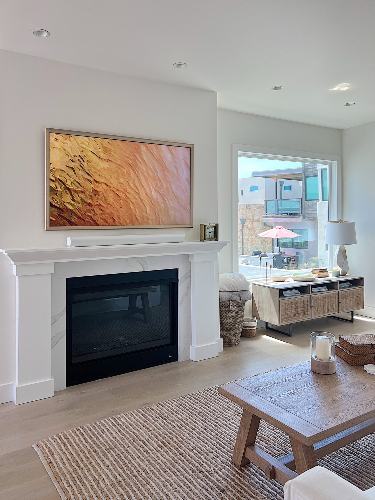 The living area at our beach house with a large fireplace, a frame tv, a window overlooking the ocean, and a couch, rug, coffee table and chair from Pottery Barn. The vintage cabinet, marble bust, and round rattan mirror are at the top of the staircase.