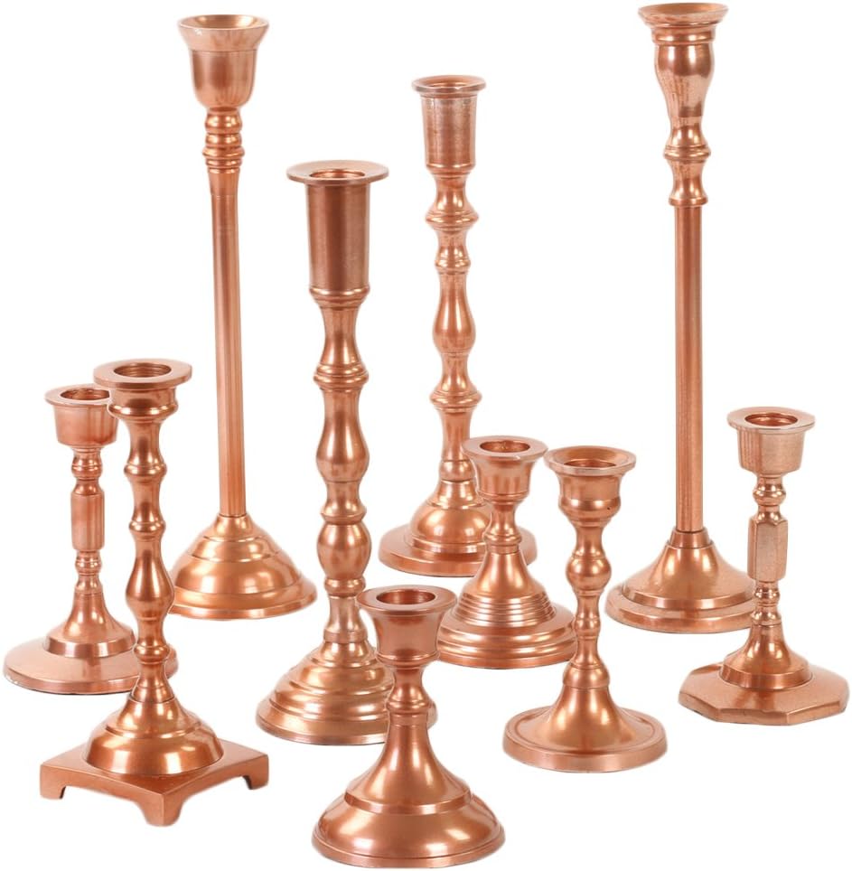 A collection of nine copper-colored metal candlesticks of varying heights and designs, arranged in a group, evokes a warm fall aesthetic, reminiscent of pumpkin spice and cozy evenings.