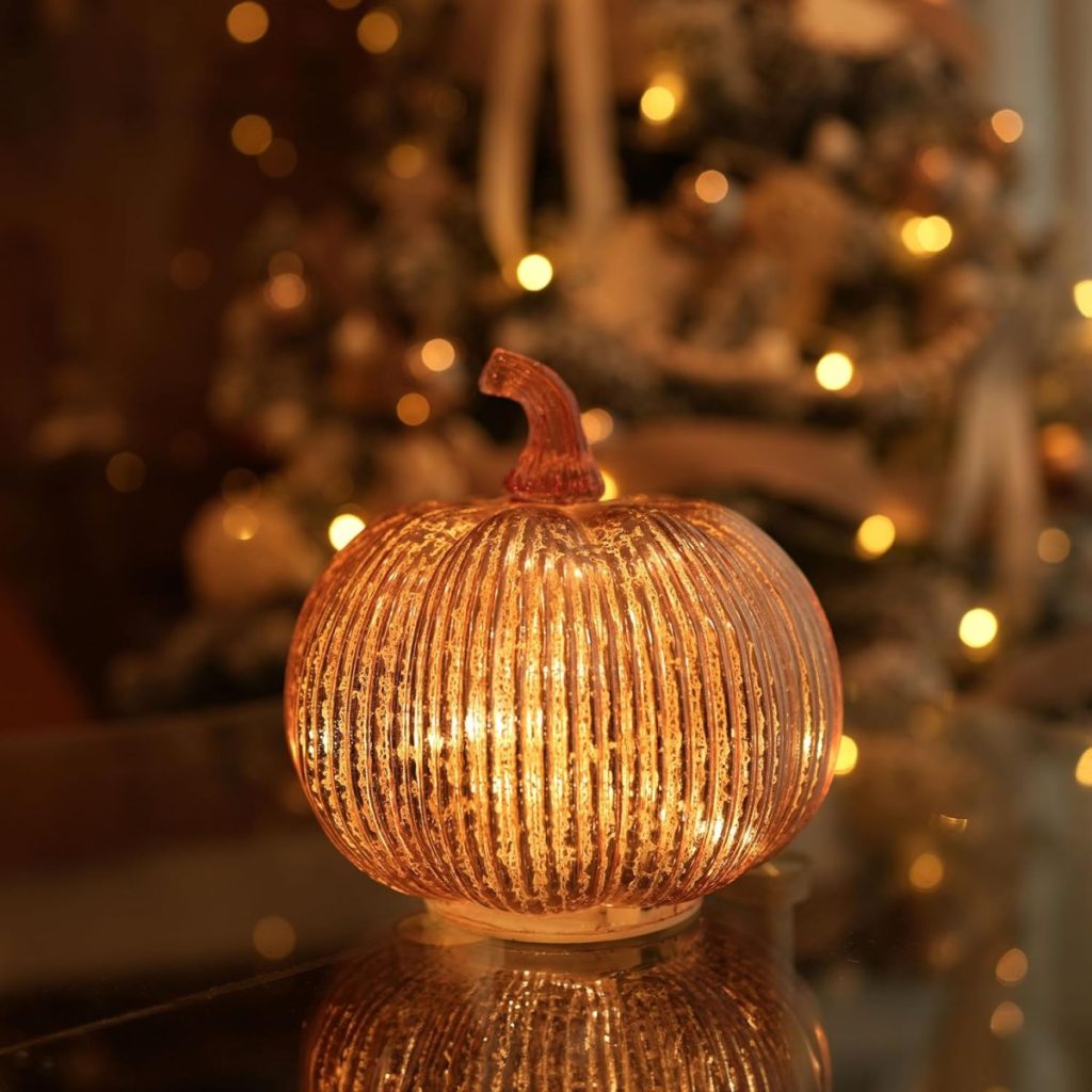 A glass fall pumpkin with internal lights sits on a reflective surface, with a blurred, illuminated Christmas tree in the background.