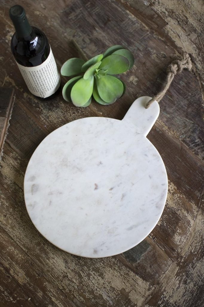 A round marble cutting board with a handle sits elegantly on a wooden surface, accompanied by a wine bottle and a small green plant, perfectly setting the stage for a delightful pear and cranberry tart.