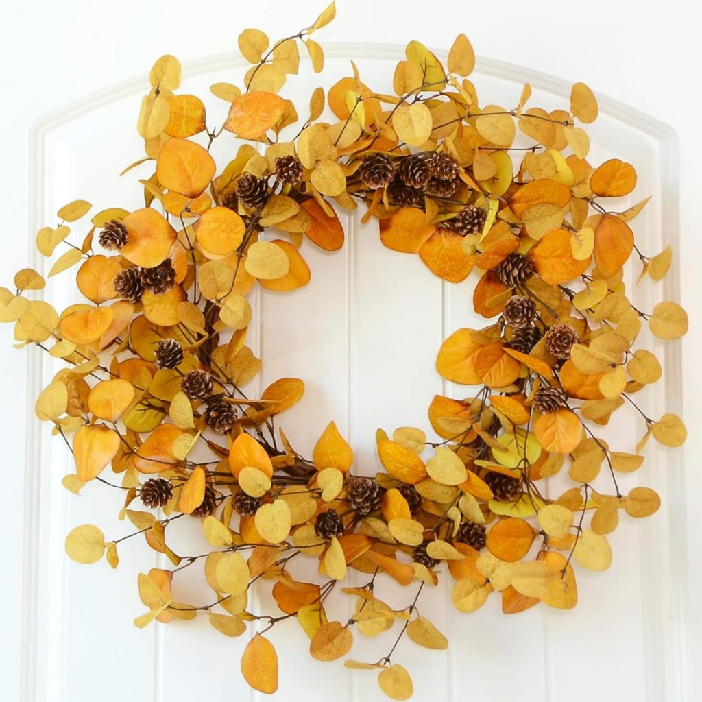 A festive Halloween wreath made of orange and yellow autumn leaves and small pinecones is hung on a white door.