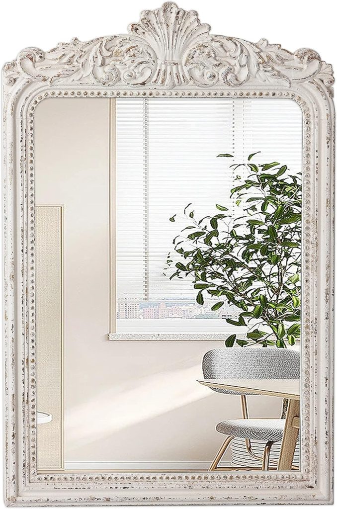 An ornate mirror with a white frame reflects a bright room featuring a plant, a cozy fall pumpkin on the windowsill, blinds, and a partially visible chair.