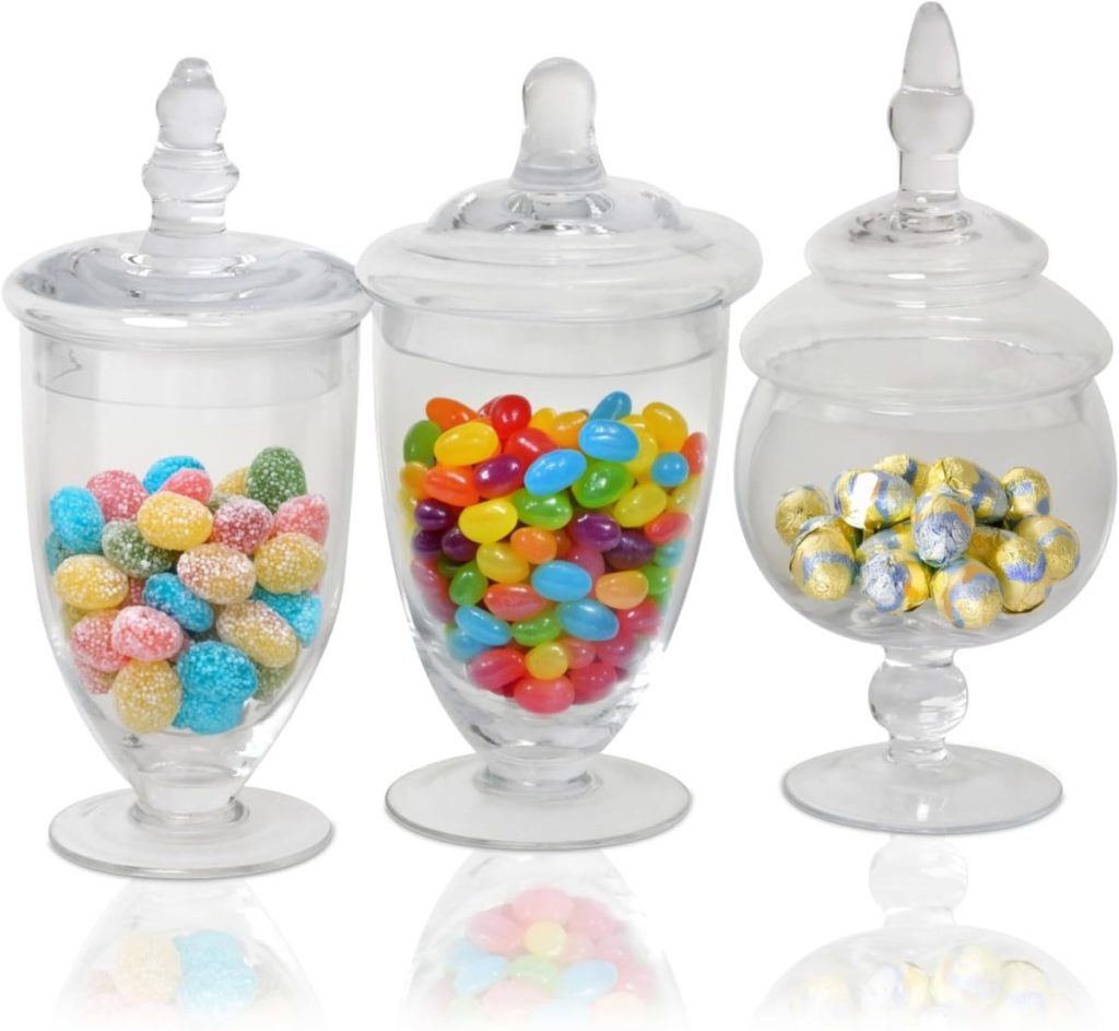 Three glass jars with lids, containing colorful candies: the left jar has blue and pink sugar-coated candies resembling Halloween sugar cookies, the middle jar has assorted jellybeans, and the right jar has gold-foil-wrapped chocolates.