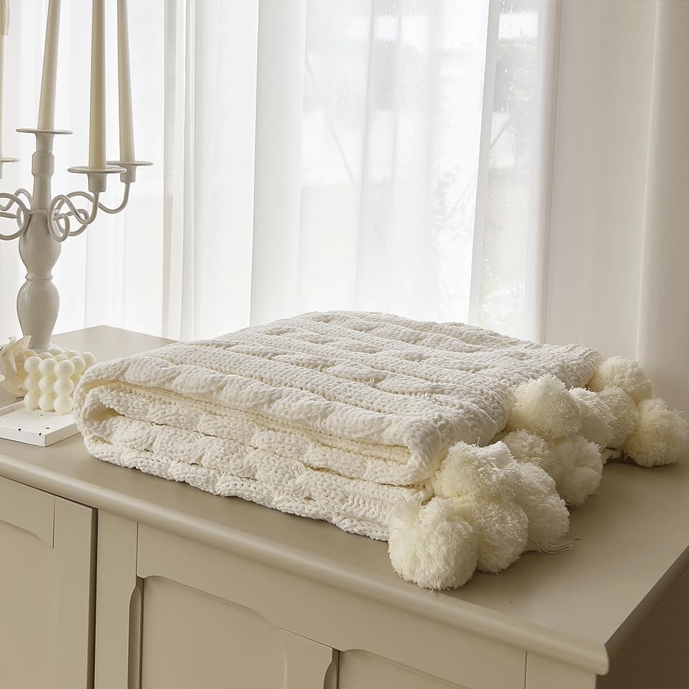 A folded cream-colored knit fall blanket with pom-poms is placed on a dresser next to a decorative candle holder in a softly lit room.