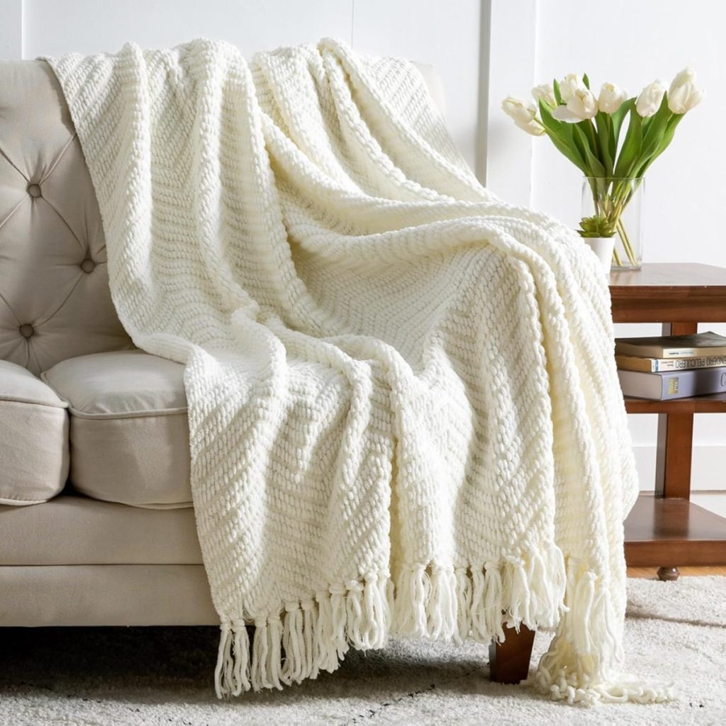 A cozy, cream-colored fall blanket with fringe draped over a beige sofa next to a wooden table and a vase of white tulips.