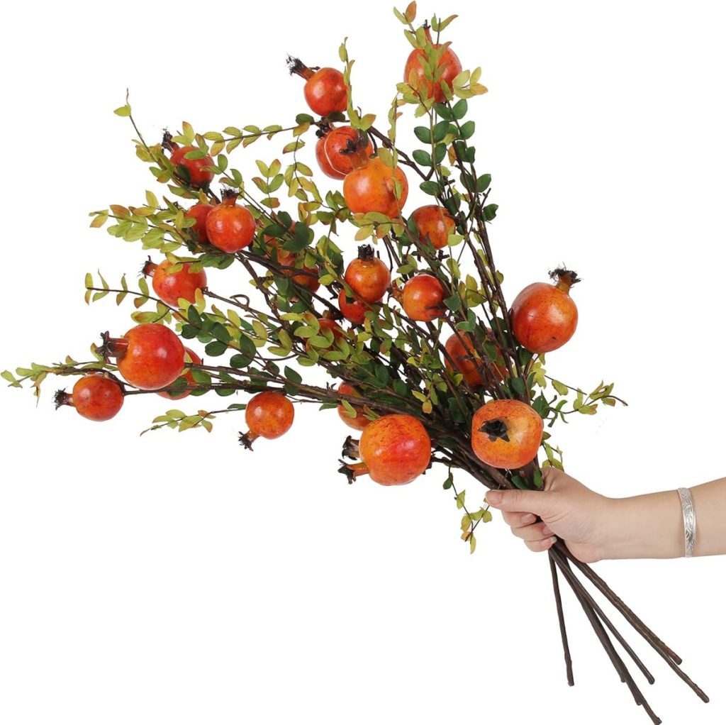 A hand holding a bouquet of artificial pomegranate branches with green leaves and red fruits, reminiscent of the vibrant hues often found in cozy fall pillows.