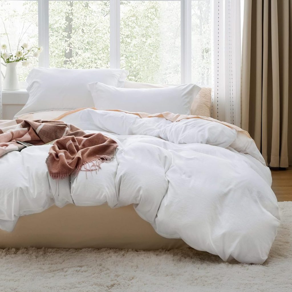 A neatly made bed with white bedding, a pink throw blanket, and pillows, sits in a bright room with large windows and beige curtains. A tray of Halloween sugar cookies adds a festive touch.