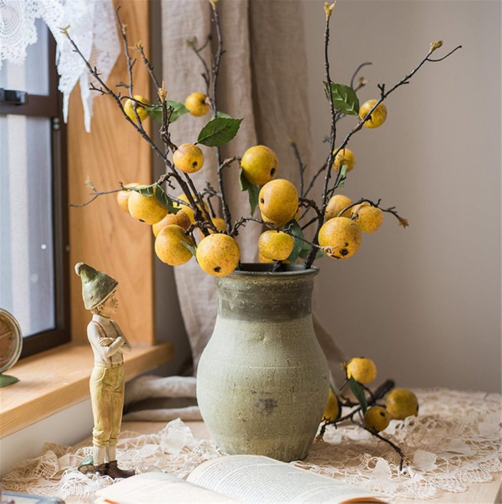 A ceramic vase holds branches with yellow fruits on a lace tablecloth, evoking the essence of a Thanksgiving wreath. A small figurine stands nearby, complemented by a window and an open book in the background.