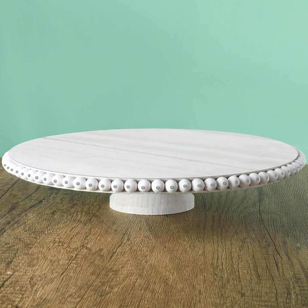 A white wooden cake stand with bead detailing on the edge sits elegantly on a wooden surface, framed by a light green background. The setting is perfect for displaying a vibrant purple sweet potato dessert.