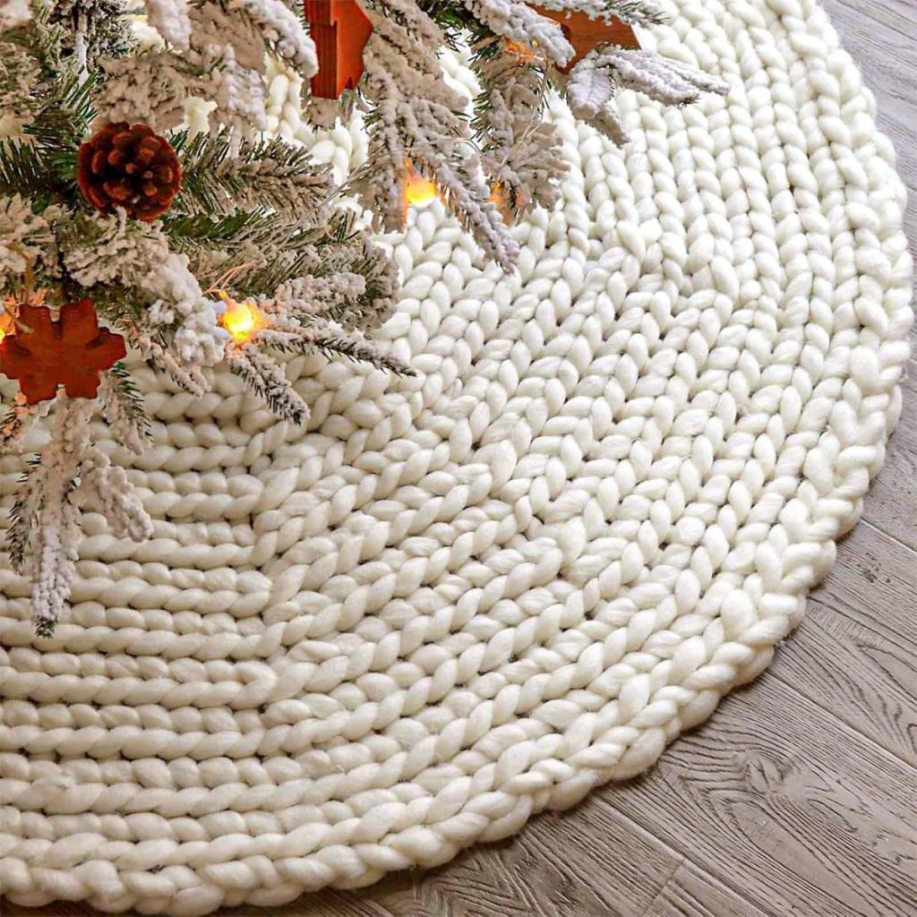 A thick, white, braided rug resembling a cozy fall blanket lies under a decorated Christmas tree adorned with lights and pinecones, set against a light-colored wooden floor.