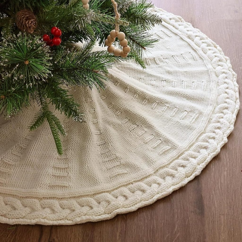 Close-up of a Christmas tree with a white knitted tree skirt on a wooden floor, resembling the cozy charm of a fall blanket.