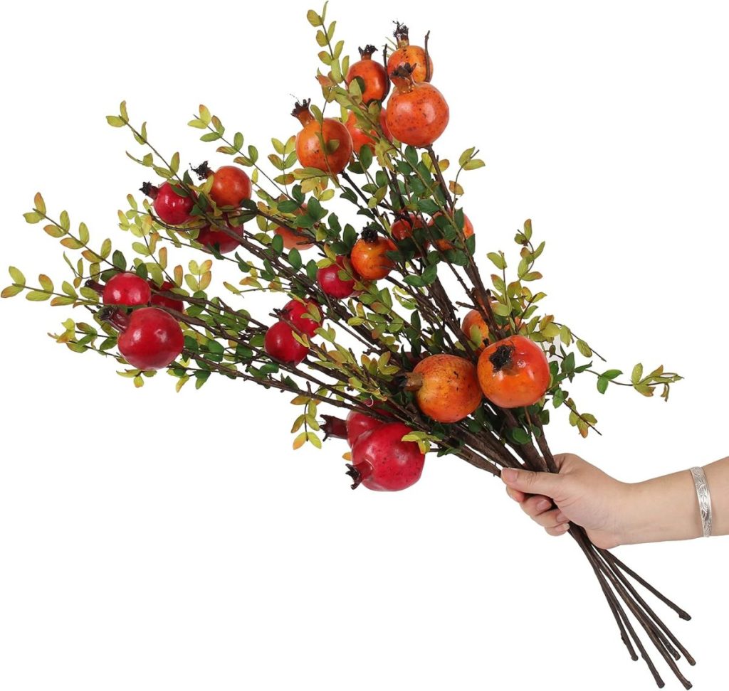 A hand holding a bouquet of branches with vibrant red pomegranates and lush green leaves evokes the cozy warmth of fall pillows against a crisp white background.