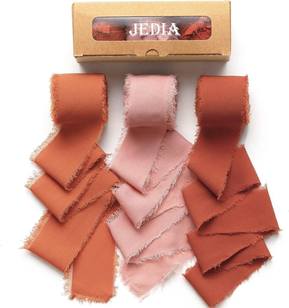 Five rolls of frayed fabric ribbons in shades of rust and pink sit ready for crafting, ideal for a Thanksgiving wreath. A cardboard box labeled "JEDIA" rests on top, adding a touch of convenience to your creative endeavor.