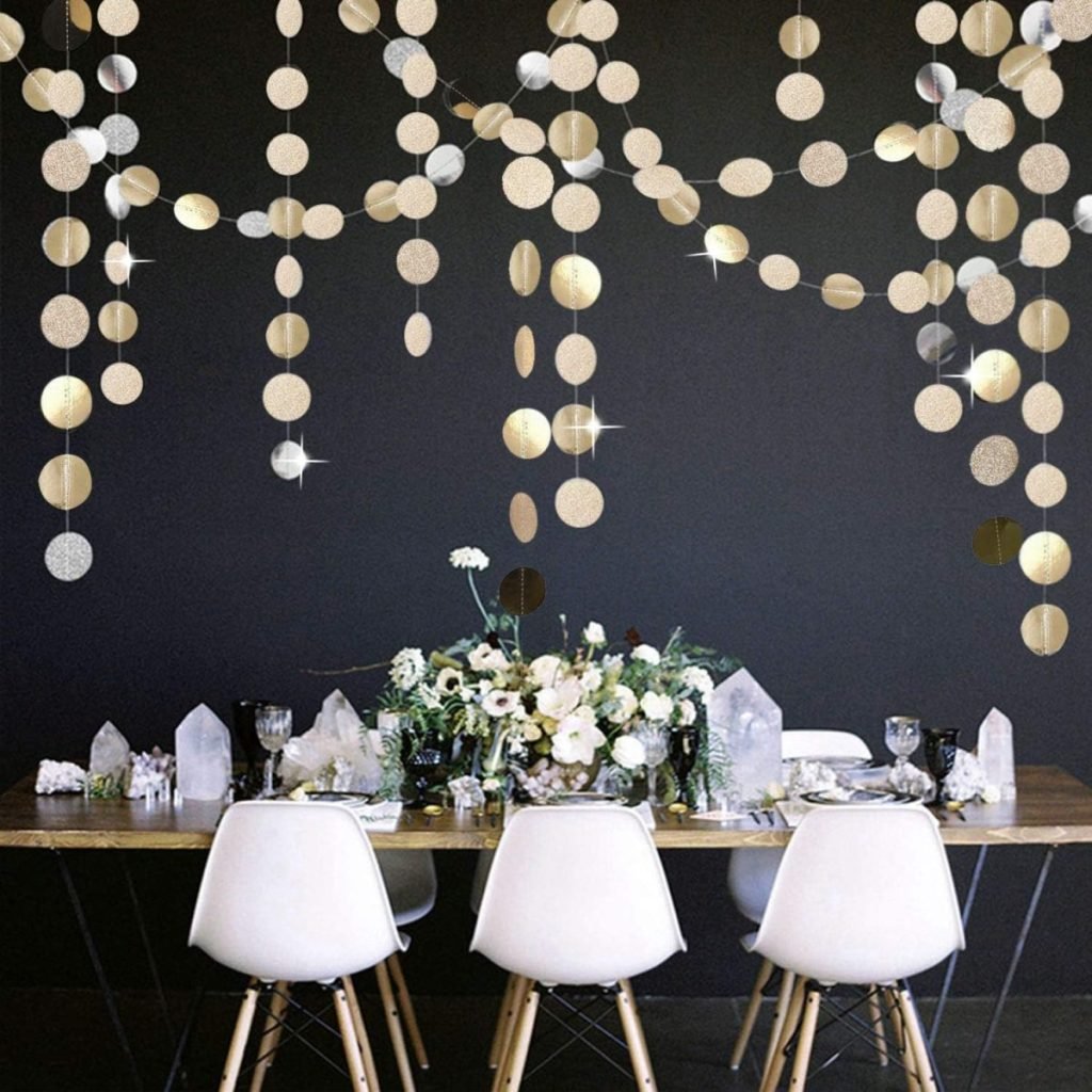 A dining table with white chairs, perfect to set a Christmas table, features a festive floral centerpiece and crystal accents. It's beautifully contrasted by a black wall adorned with hanging gold and silver circular garlands.