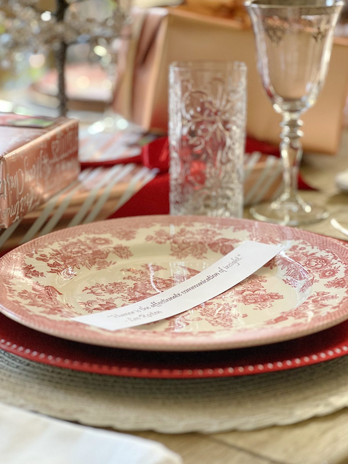 A Christmas table set in cranberry and rose gold colors with a DIY tree, cranberry and white plates, amber and clear glasses, rose gold chargers, and wrapped packages.