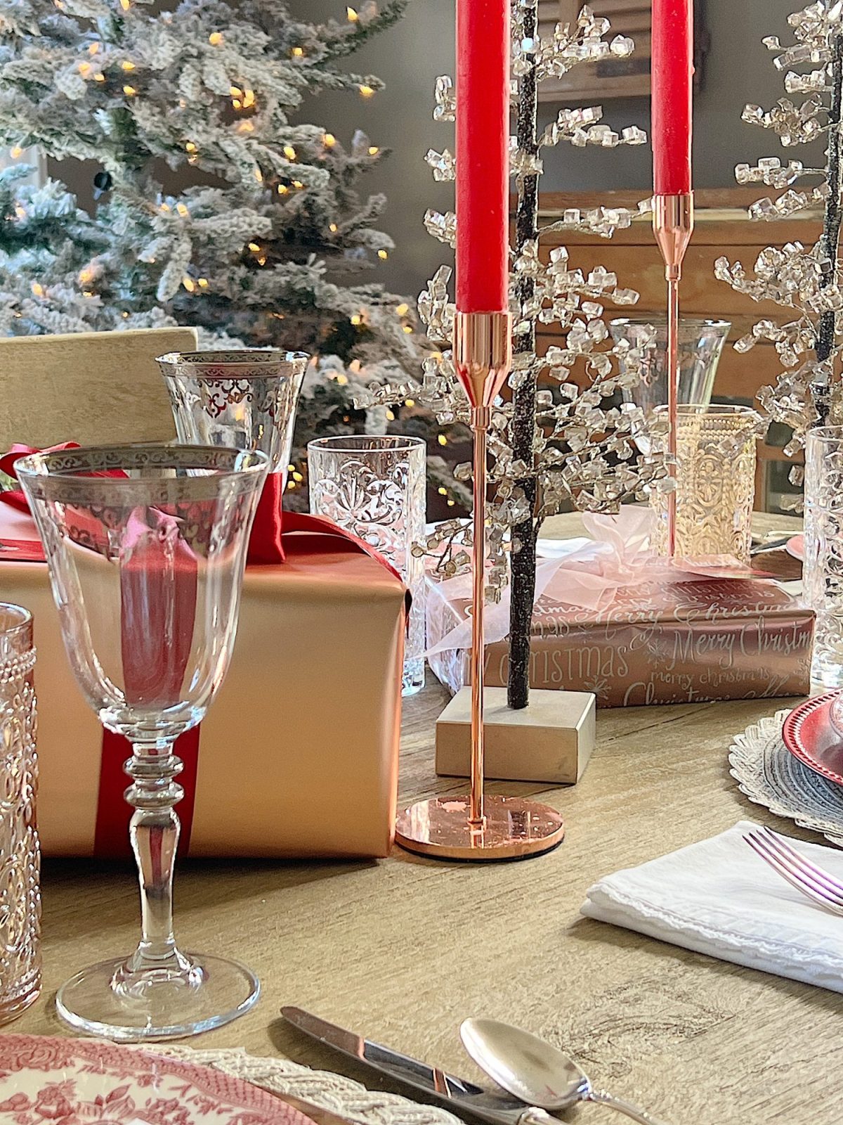 A Christmas table set in cranberry and rose gold colors with a DIY tree, cranberry and white plates, amber and clear glasses, rose gold chargers, and wrapped packages.
