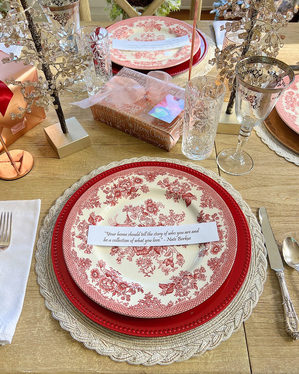 A Christmas table set in cranberry and rose gold colors with a DIY tree, cranberry and white plates, amber and clear glasses, rose gold chargers, and wrapped packages.