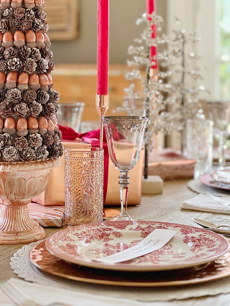 A Christmas table set in cranberry and rose gold colors with a DIY tree, cranberry and white plates, amber and clear glasses, rose gold chargers, and wrapped packages.