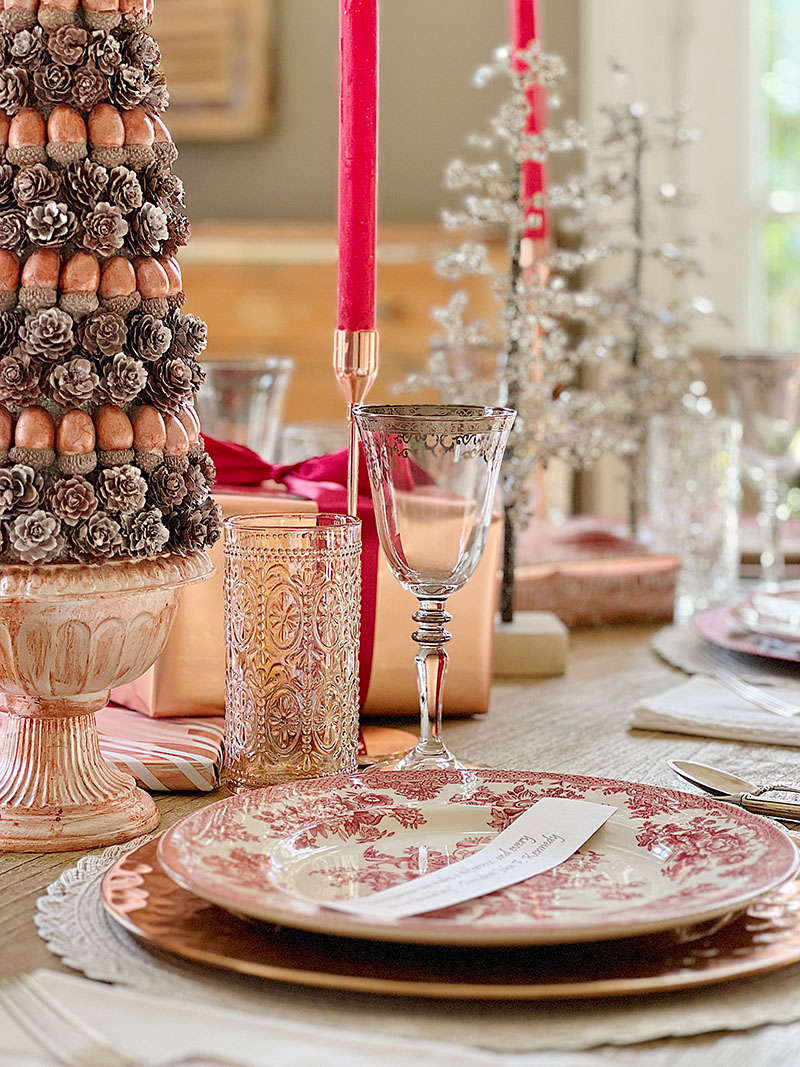A Christmas table set in cranberry and rose gold colors with a DIY tree, cranberry and white plates, amber and clear glasses, rose gold chargers, and wrapped packages.